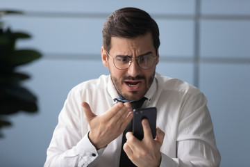 Frustrated businessman angry over bad news message on smartphone. Unhappy man employee argue using...