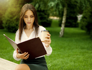 Fototapeta premium Young cheerful woman manager holding a folder with documents in her hands and drink coffee while sitting on bench in park. Business concept