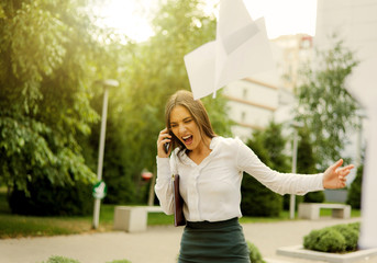 Brought to hysteria, angry business woman dressed in a white blouse and skirt, screaming in the phone, throws out sheets of paper outdoors in the city