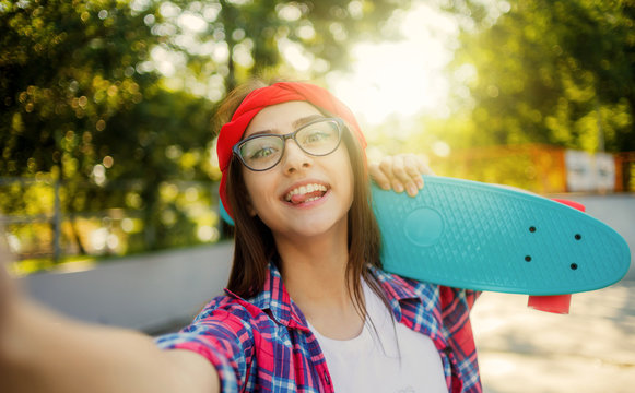 Selfie portrait of funny young woman in glasses and red plaid shirt. Hipster girl fooling around in skatepark while taking picture with skateboard in skatepark at sunny bright day.