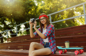 Funny female skater dressed in stylish clothes sits in the stands in skatepark while holds retro camera and listens to music in earphones at bright sunny day © splitov27