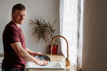 A man of European appearance in a burgundy T-shirt washes dishes in the kitchen by the window