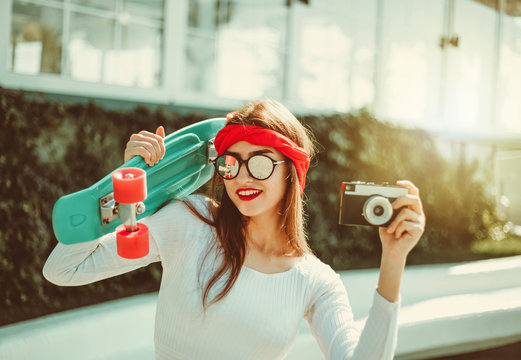 Retro style. 80s. Portrait of stylish dressed young woman with skateboard and film camera outdoors on bright sunny day. Summer lifestyle image of trendy pretty young girl
