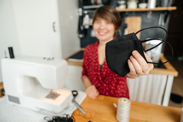 Young Woman shows a black protective mask that he made on his own at home in the kitchen. Home production. Remote work.