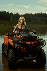 Beautiful blonde girl on a ATV rides on water and sand mountains. Green forest all around. In summer season - 343034717