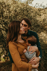 Couple standing with their son outdoors. Young family concept. Mother holds the baby in the arms. Man kiss the woman in the cheek.