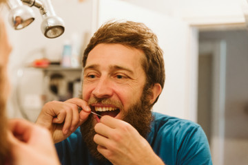 Handsome bearded man looking at mirror while using dental floss in bathroom 