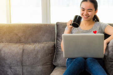 Beautiful Asian woman holds a coffee cup smile and look at the laptop at work online, Working remotely with a computer for business on the sofa in the home, Cute girl communication sunlight background
