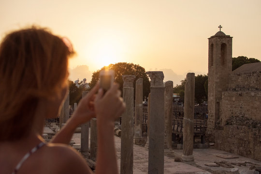 Happy young woman takes pictures on smartphone of the ancient temple at sunset. Paphos, Cyprus