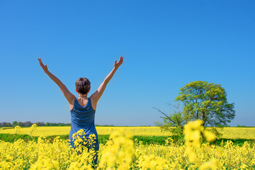 Naklejka na ściany i meble Happy woman wearing blue top and jeans raised her hands high against blu sky and yellow rapeseed fields