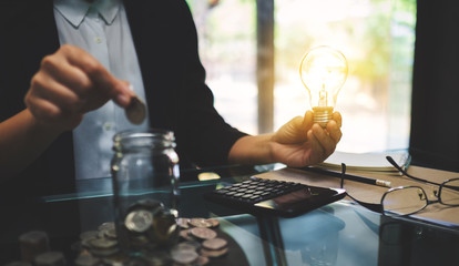 Businesswoman holding a light bulb while putting coin into a glass jar for saving energy and money concept