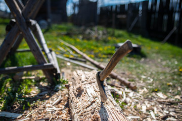 Axe with wood handle in a pine log, wood chips on the ground, wooden sawhorse, shallow depth of field, focus on the  handle and the tip of the blade.