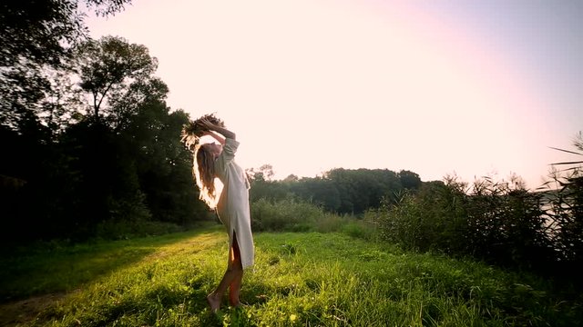 young blonde girl at sunset in a vintage linen dress puts a wreath of flowers on her head. sun rays shine on the background, green grass forest and nature landscape. Woman with long hair