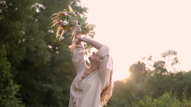 young blonde girl at sunset in a vintage linen dress puts a wreath of flowers on her head. sun rays shine on the background, green grass forest and nature landscape. Woman with long hair