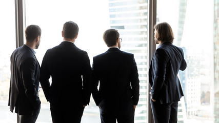 Rear view close up four confident successful businessmen looking through large panoramic window,...
