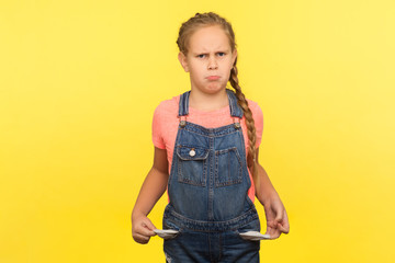 Portrait of upset little girl with braid in denim overalls turning out empty pockets, having no...