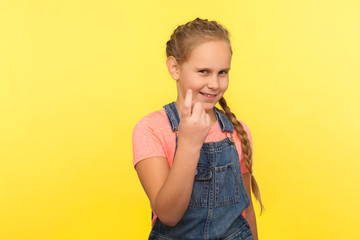Approach to me! Portrait of cheerful cunning little girl with braid in denim overalls gesturing come here and smiling slyly, inviting with beckoning finger. studio shot isolated on yellow background