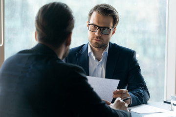 Serious attentive HR manager wearing glasses looking at job applicant, holding cv, listening to...