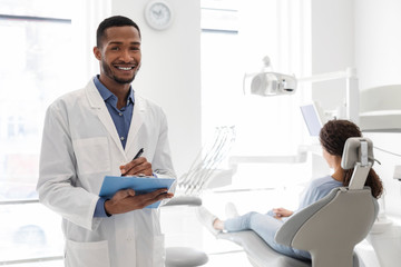 Black dentist smiling and holding folder in dental cabinet