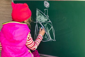 A girl in a pink jacket draws with chalk on a school blackboard a house. In her hands is chalk