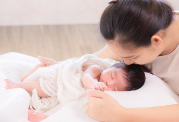 Selective Focus, Asian mother kiss adorable newborn while sleep, mom support baby girl with love, care, protection hold hand together. Motherhood lifestyle take care infant on bed at home concept