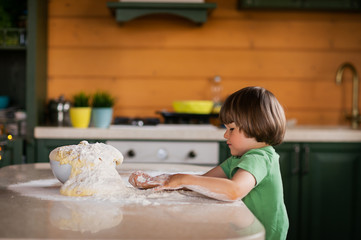 Fototapeta na wymiar Little boy with beautiful dark hair in green t-shirt kneading the dough and cooking pizza