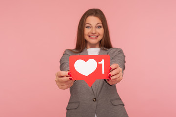 Popular content, social media trends. Positive elegant woman in business suit holding network heart Like icon and smiling to camera, recommending to follow blog. indoor studio shot, pink background