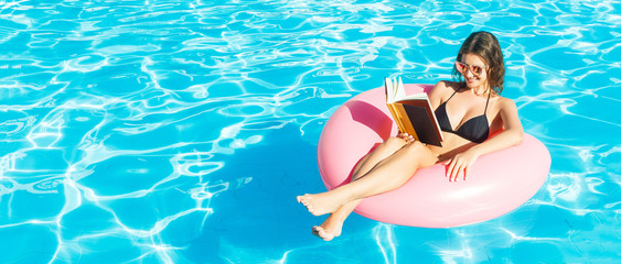 Young woman is reading a book sitting on the inflatable ring in the swimming pool. Summer...