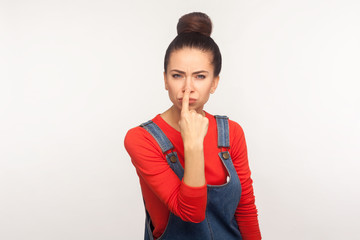 Portrait of girl with hair bun in overalls pointing nose doing lie gesture, suspecting trickster in...