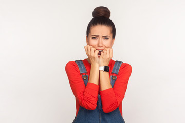 Portrait of depressed nervous girl with hair bun in denim overalls biting nails and looking with scared eyes, anxious terrified expression, neurotic person. studio shot isolated on white background