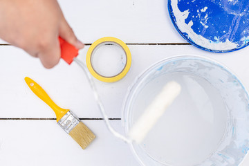 Painting brush in the hand and paint bucket on the white wooden table with painting brushes and tape. Painting and DIY concept with painting hand tools with paint in the bucket