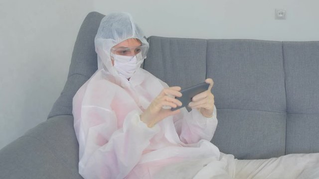 Woman in protective suit is browsing smartphone on coronavirus pandemic quarantine. Hypochondriac at home sitting on couch. COVID-19 epidemic, 2019-ncov pandemic. Fear of infection.