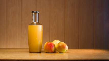 Big glass bottle with apple juice and three apples on a wooden background with copy space.