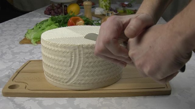 Cutting a large head of fresh homemade goat cheese. Textured surface of white cheese lying on the warm surface of a wooden board with fresh vegetables in the background.