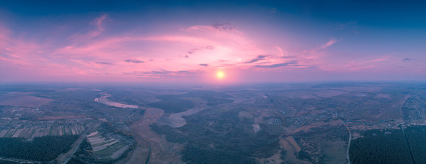 Spring rural landscape in the evening with a beautiful burning sky. Aerial view. Panoramic view of pine forest, fields, and river during blazing sunset. Panorama 180 from 21 images