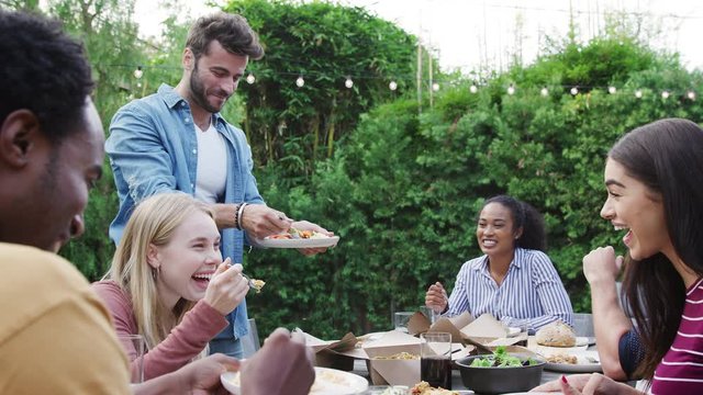 Multi-Cultural Friends At Home Sitting At Table Enjoying Food At Summer Garden Party