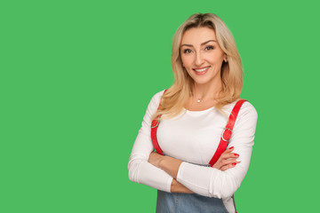 Portrait of positive self assured beautiful adult woman in stylish overalls holding arms crossed, looking at camera with toothy smile and confident expression. studio shot isolated on green background