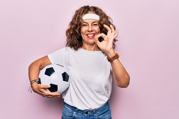 Middle age beautiful sporty woman playing soccer holding football bal over pink background doing ok sign with fingers, smiling friendly gesturing excellent symbol