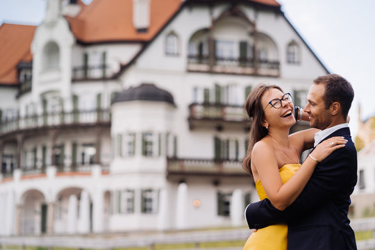 Romantic Couple In Love Hugging  Against An Old Building. Young Woman Dressed In Yellow Evening Dress, Laughing Happily She Get A Good News. Happy People Concept