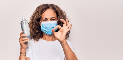 Middle age woman wearing coronavirus protection mask holding hand sanitizer bottle doing ok sign with fingers, smiling friendly gesturing excellent symbol