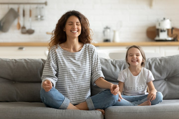 Full length happy young nanny mom sitting on sofa in lotus pose, teaching small daughter yoga...