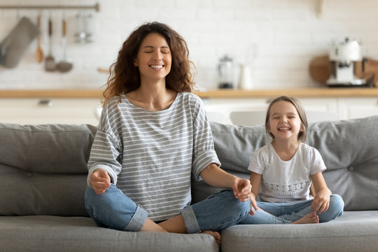Full length happy young nanny mom sitting on sofa in lotus pose, teaching small daughter yoga breathing exercise. Friendly sincere two female generations family relaxing on couch, healthcare concept.