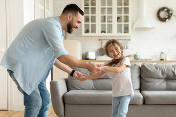Smiling single dad enjoying funny family activity with laughing little preschool daughter in modern studio living room. Happy small child girl dancing to favorite pop music with joyful father indoors.