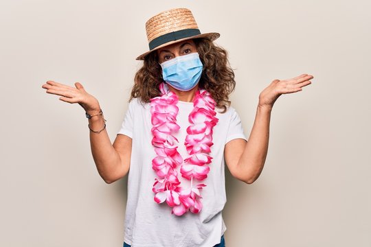 Middle age tourist woman wearing coronavirus protection mask and summer hat on vacation clueless and confused with open arms, no idea and doubtful face.