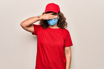 Middle age delivery woman wearing red cap and coronavirus protection mask peeking in shock covering face and eyes with hand, looking through fingers afraid