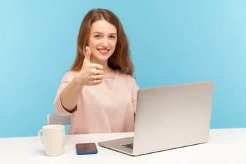 Excellent job! Positive satisfied woman with charming toothy smile showing thumbs up gesture, like, approval sign, working on laptop from home office. indoor studio shot isolated on blue background