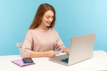 Internet shopping, online utility payments. Positive smiling woman holding credit card and typing on laptop, making purchase or service, using computer. indoor studio shot isolated on blue background