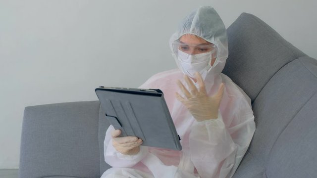 Woman hypochondriac in protective suit, mask, glasses, gloves talking video chat on tablet sitting on couch at home in coronavirus quarantine on pandemic. COVID-19 epidemic, 2019-ncov pandemic.