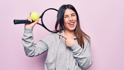 Young beautiful sportswoman playing tennis using racket and ball over pink background smiling happy pointing with hand and finger