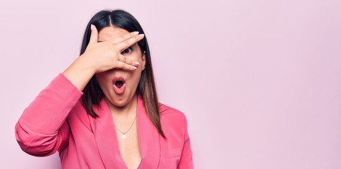 Young beautiful business woman wearing elegant jacket standing over pink background peeking in shock covering face and eyes with hand, looking through fingers afraid